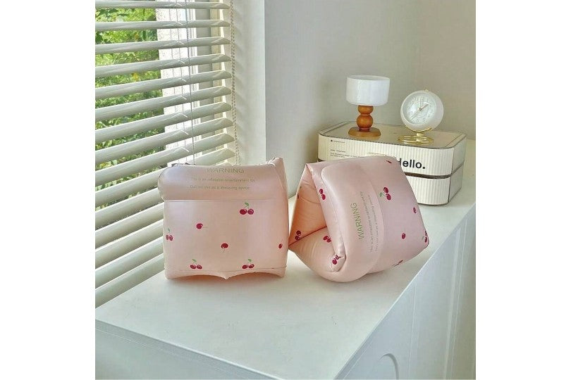 Two pink inflatable arm floats with cherry design on a white surface near a window with blinds.